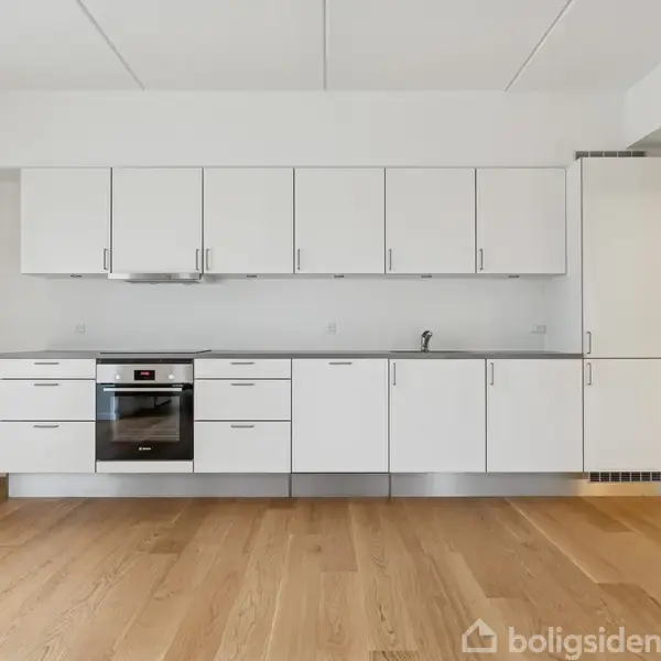 Hvidt køkken med moderne skabe og indbygningsovn, i et rum med trægulv. Køkkenet har en vask integreret i bordpladen, omgivet af en minimalistisk og lys indretning.