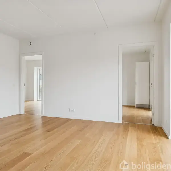 Et tomt rum med lyst trægulv viser flere døråbninger mod andre værelser. Hvide vægge og store vinduer giver lys og rummelighed.