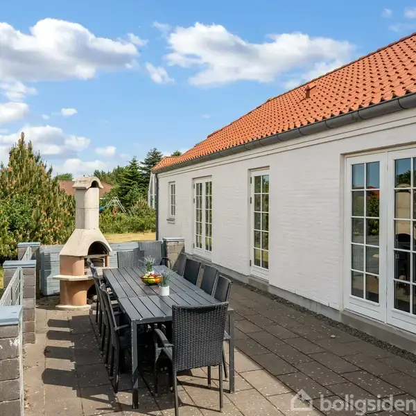 Udendørs terrasse med havemøbler og grillpejs ved siden af et hvidt hus med rødt tag. Omgivet af grønne træer under en blå himmel med skyer.