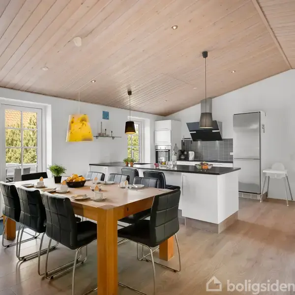 Et spisebord med sorte stole er dækket til morgenmad. I baggrunden ses et moderne køkken med hvidt interiør. Udendørs terrasse synlig gennem store glasdøre.