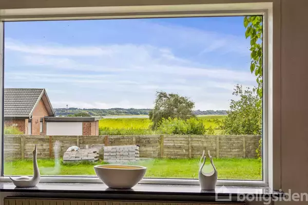 En vindueskarm med dekorative genstande, såsom en skål og figurer, har udsigt til en have med grønne marker og en garage i baggrunden under en blå himmel.