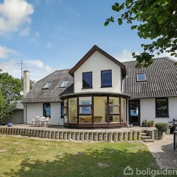Et hvidt hus med grå tagsten har en rund glaskarnap. Huset står på en stor græsplæne omgivet af træer under en blå og skyet himmel.