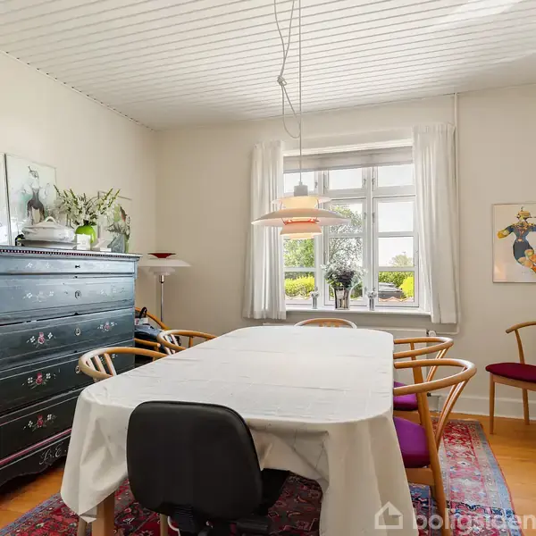 Et spisebord dækket med hvid dug står i midten af et lyst rum. Omkring bordet er der stole, og en kommode står langs væggen. Et vindue med gardiner giver udsyn til haven.