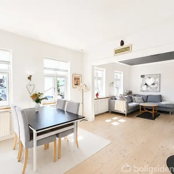 Stue med to rum i lyse toner. Et spisebord med stole står ved vinduerne, mens en sofa er placeret i hjørnet. En brændeovn står i forgrunden.