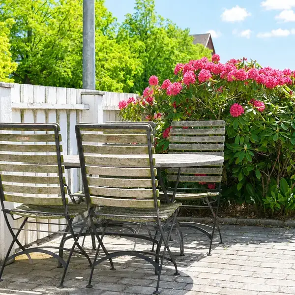 Tre havestole og et bord står på en flisebelagt terrasse ved siden af en hvid hegn. Lyserøde blomster blomstrer i en busk til højre. Himmelen er blå med få skyer.