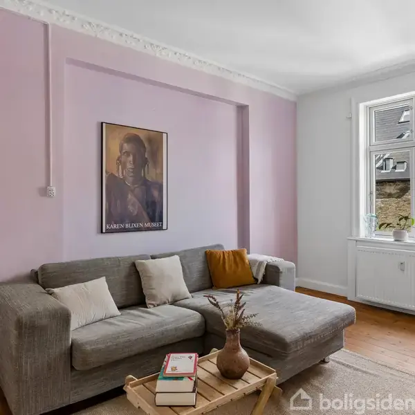Grå sofa med puder står mod en lys lilla væg med en plakat af Karen Blixen hængende. Foran sofaen står et sofabord med bøger og en vase. Vindue til højre.