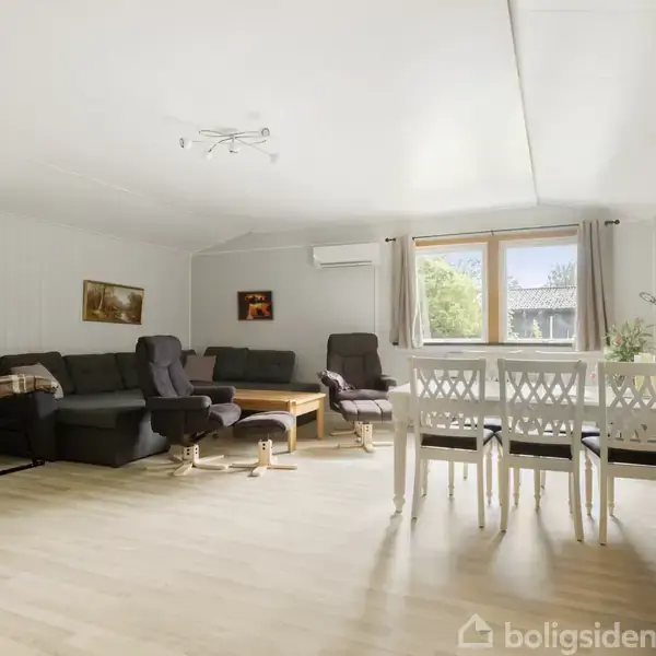 Stue med gråt hjørnesofaarrangement og to lænestole omkring et sofabord til venstre; omkring et spisebord med fire stole til højre. Store vinduer med gardiner.