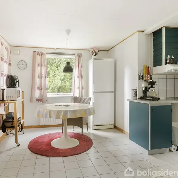 Køkken med rundt bord på rødt tæppe, omgivet af hvide fliser og blå skabe. Et vindue med lyserøde gardiner og en pendellampe giver lysindfald.