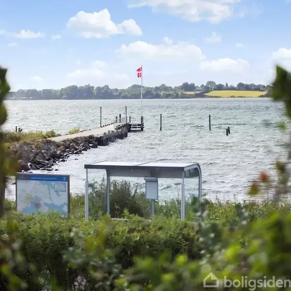 Et busskur står stille nær en kyst, omgivet af buske. I baggrunden er der en mole med et dansk flag, og havet strækker sig under en skyet himmel.