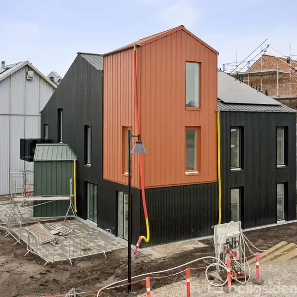 Moderne hus med blandet facade i sort og rød-orange paneler. Ved siden af pågår byggearbejde med stilladser. Omgivet af bar jord og byggepladsudstyr. Ingen tekst fundet.