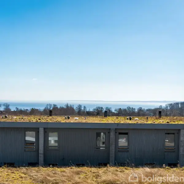 Bygning med fladt grønt tag står stille i forgrunden. I baggrunden ses en vid udsigt over havet under en blå himmel, omkranset af træer.