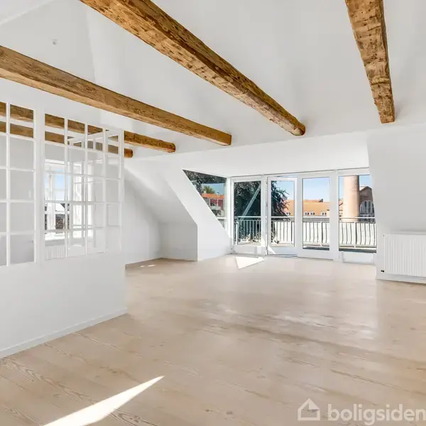 Et lyst og rummeligt loftsværelse med trægulv og fritlagte bjælker, store vinduer til venstre og en glasvæg til højre, giver udsigt til en solrig balkon udenfor.