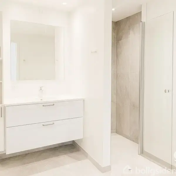 Et moderne badeværelse med en hvid vaskeskab, en spejl over skabet, og en flisebelagt bruseniche. Rummet er lyst og enkelt indrettet.