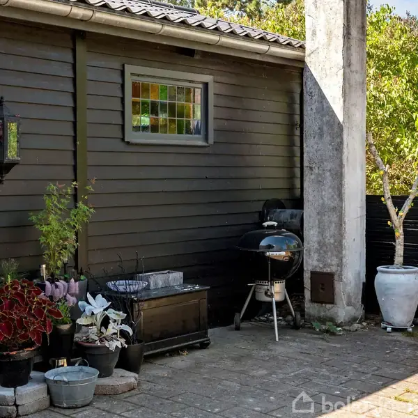 Grill står på terrasse ved siden af krukker med planter. En træbænk er placeret tæt på. Træbeklædt væg har farvet glasvindue og lanterner. Omgivelserne er grønne og solrige.