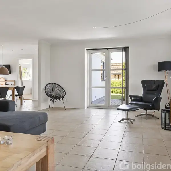 Stue med lænestol ved glasdøre, der åbner mod en terrasse. Sofa og sofabord til venstre; spisebord og stole i baggrunden. Lyse fliser og en minimalistisk stil.