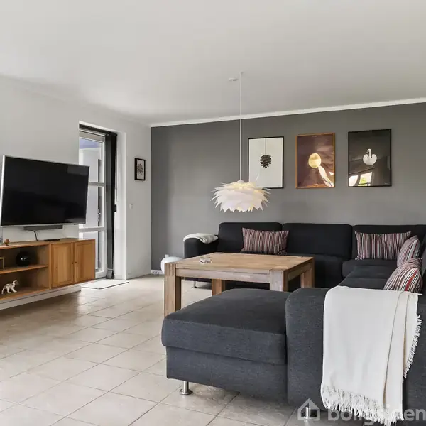 En moderne stue indeholder en mørk hjørnesofa med puder, på væggen hænger tre billeder. Et træbord står foran sofaen. Til venstre ses et TV på et lavt skab.