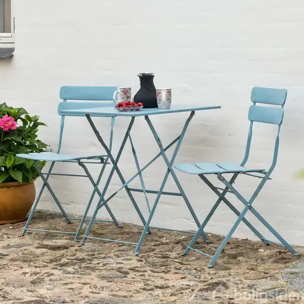 Blå bord og stole står på stenet gårdhave. Bordet har kaffekande, kopper og jordbær. En potte med lyserøde blomster er ved siden af, op ad hvid murstensvæg.