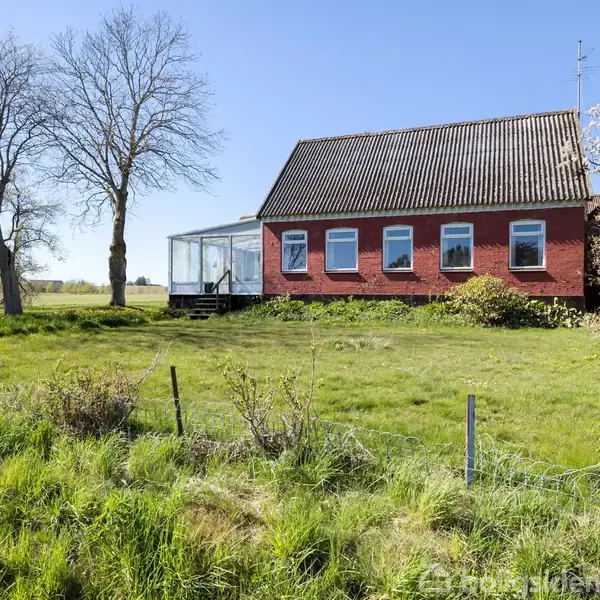 Et rødt murstenshus står alene på en græsmark. Huset har flere vinduer og en glasterrasse. Tre bladløse træer står omkring huset under en klar blå himmel.