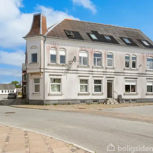 En hvid, to-etagers bygning med adskillige vinduer står på et gadehjørne. Fortovet omgiver bygningen, og himlen er klar med nogle få skyer. Ingen tekst er synlig bortset fra "Boligsiden" vandmærket.