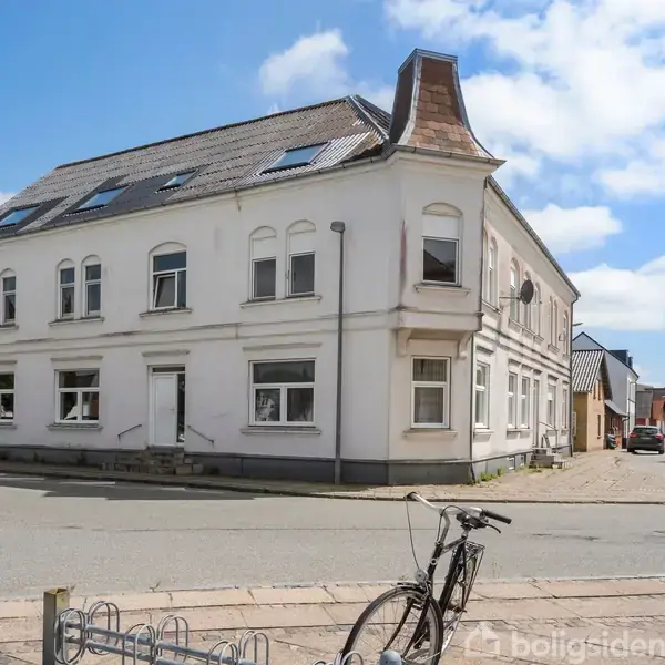 En hvid, to-etagers bygning med skifertag står på et gadehjørne. En cykel er parkeret foran bygningen. Gaden er tom under en blå himmel med få skyer.