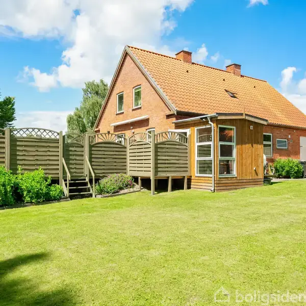 Et rødt murstenshus med et orange tag står stille i et solrigt boligområde. Græsplæne og grønne buske omgiver huset. Der er et træhegn langs grunden.