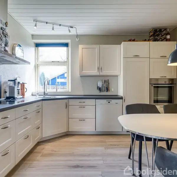 Et lyst køkken med hvide skabe og en sort bordplade. Der er en rund spisebord med fire stole. En hængelampe lyser området op, og et vindue findes i baggrunden.