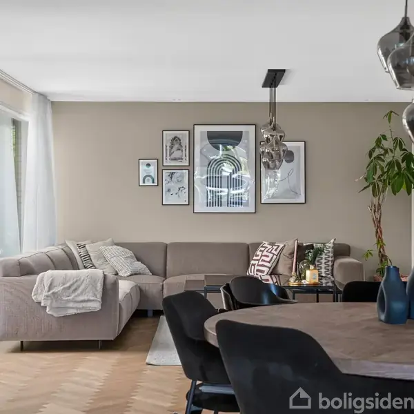 En beige hjørnesofa står i en stue med en væg fyldt med kunst. Spisebord med stole og dekorative lamper i forgrunden. Store vinduer giver lysindfald. Ingen tekst.