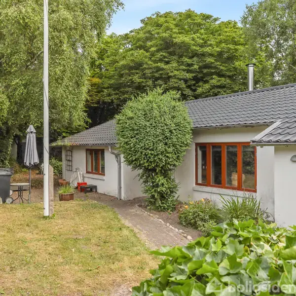 Et hvidt hus med sort tag står fredfyldt i en grøn have med høje træer. Nummeret 22 er synligt ved indgangen, og en terrasse med parasol ses til venstre.