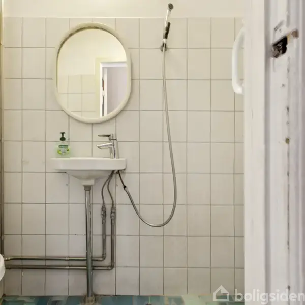 Et rundt spejl hænger over en vask med en flydende sæbedispenser. En håndbruser er placeret på en holder. Omkring er der hvide fliser i et badeværelse.