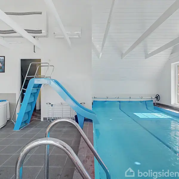 En swimmingpool med en blå rutsjebane ved siden af. I baggrunden ses et spabad. Rummet er lyst med hvide vægge og et stort vindue.