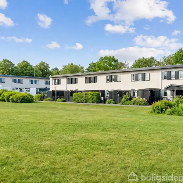 Rækkehus med flad tag og store vinduer, beliggende på en række med lignende huse. En lille have foran og en fælles baggrund af træer og blå himmel.