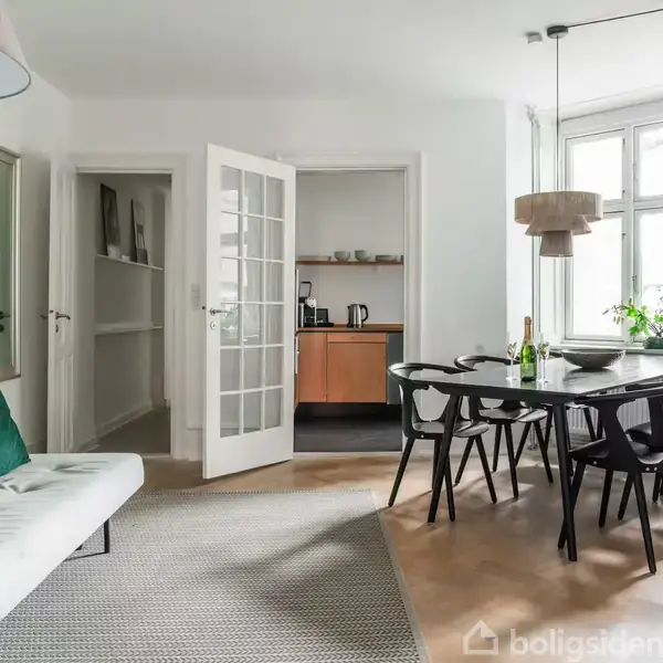 En gårdhave med træbænke omgivet af beplantning og pergolaer. Høje, hvide lejlighedsbygninger danner en rektangulær ramme omkring området, mens træer og parasoller tilføjer grønt og skygge.