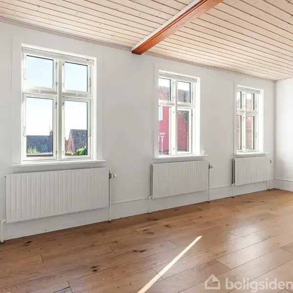 Et lyst rum med trægulv og tre vinduer. Vinduerne giver naturligt lys og udsigt til bygninger udenfor. Væggene er hvide, og der er radiatorer under hvert vindue.