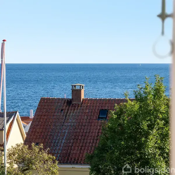Røde tage og en hvid flagstang ses foran en rolig blå havoverflade; grønne træer omkranser husene, set gennem et vindue i forgrunden.