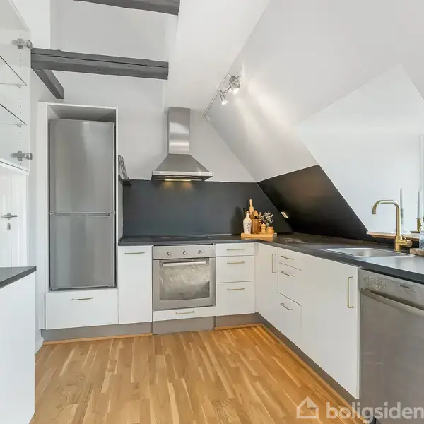 Et moderne køkken med hvidt interiør, hvor komfur, opvaskemaskine og køleskab er integreret i skabene. Køkkenet har trægulv, mørk bordplade, og skråvægge med indbygget belysning.