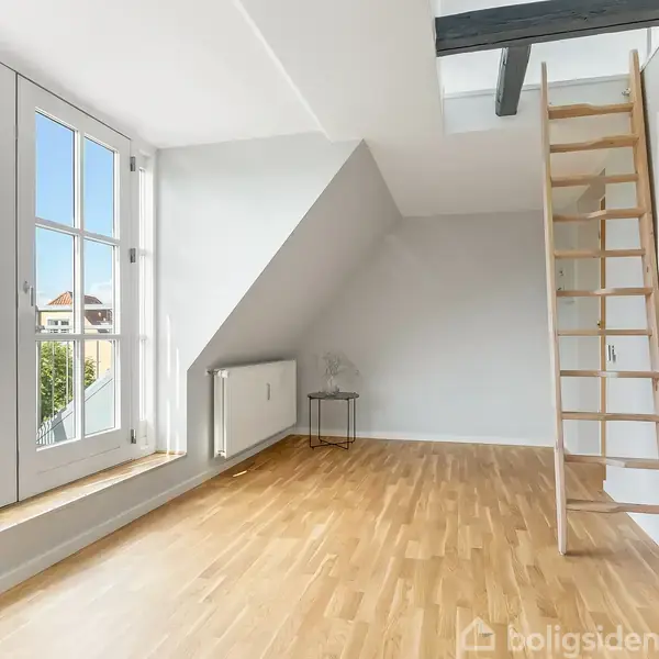 Et lyst rum med trægulv indeholder en trætrappe, der fører til en hems. Store vinduer giver udsigt til grønne omgivelser, og der ses en radiator og et lille bord.