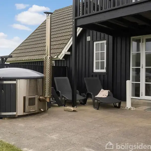 En terrasse indeholder et rundt, træfyret vildmarksbad ved siden af to sorte liggestole. Det hele er omkranset af et sort træhus med store glasdøre og vinduer, under en blå himmel.
