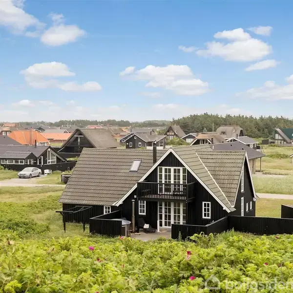 Et sort træhus med hvide vinduesrammer står i et grønt, åbent landskab. Flere lignende huse kan ses i baggrunden under en blå himmel med skyer.