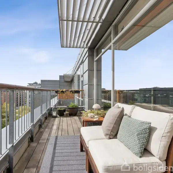 Sofasæt med puder står på altan, omgivet af glasvægge og trægelænder. Udsigten viser moderne bygninger og grønne områder under en blå himmel.