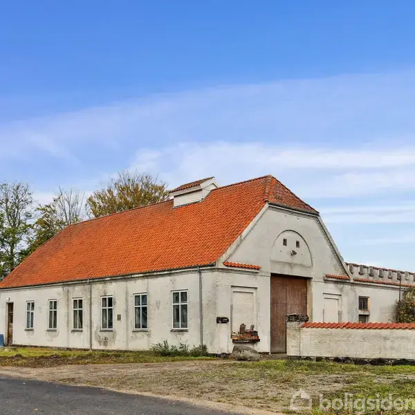 En hvid bygning med rødt tegltag står ved en vej. En høj, rød murstensskorsten strækker sig opad. Omgivet af træer og en åben himmel.