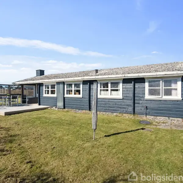 Et sort træhus står stille på en grøn plæne under en klar blå himmel, med en overdækket terrasse til venstre og en parasol i midten af græsplænen.