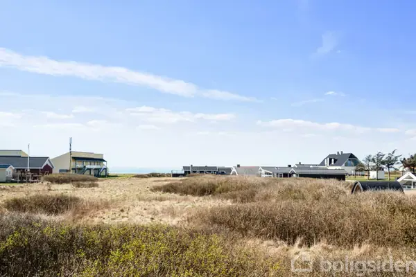 Feriehuse står stille i en kystnær landsby med klitter og græs i forgrunden under en blå himmel.