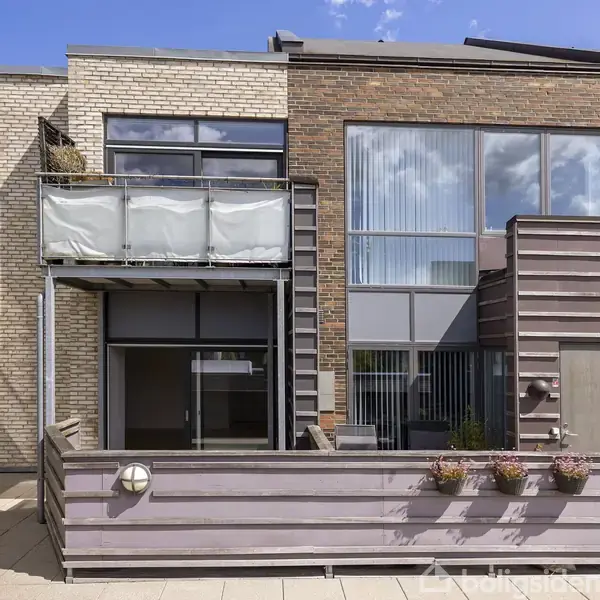 Et moderne rækkehus med store vinduespartier og en altan med gennemsigtigt gelænder. Foran er der en terrasse med blomsterkasser. Huset har murstenfacade og ligger i et nabolag.