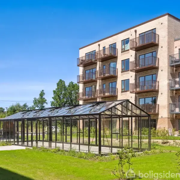En fem-etagers moderne bygning med altaner står ved siden af et drivhus i en velholdt grøn have. Skyfri blå himmel danner baggrunden.