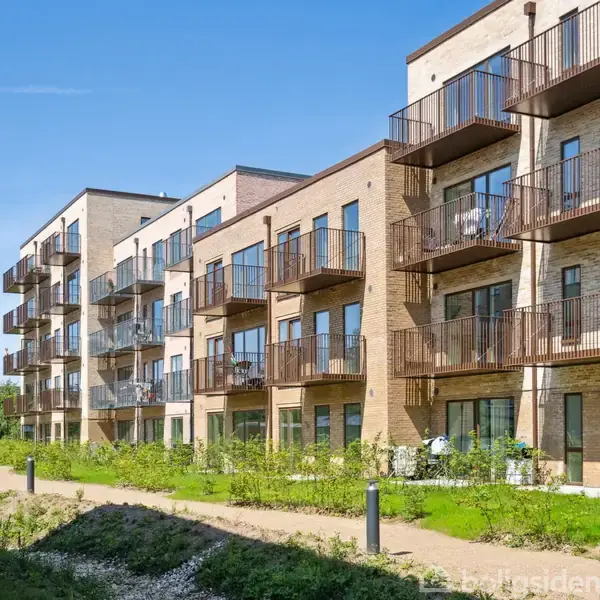 Lejlighedskompleks med flere etager, hvor altaner strækker sig ud fra hver lejlighed. Bygningen er omgivet af en velholdt græsplæne og stier under en klar blå himmel.
