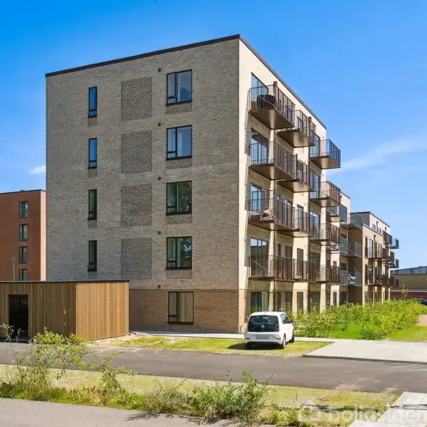 Et fem-etagers lejlighedskompleks i moderne stil står i et grønt boligområde. Flere altaner pryder bygningens facader. En parkeret hvid varebil ses i forgrunden.