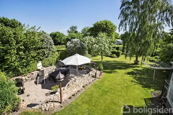 En udendørs terrasse med parasol og havemøbler står på en stenbelagt plads. Omgivelserne består af en velplejet græsplæne med træer og buske i solskinsvejr.