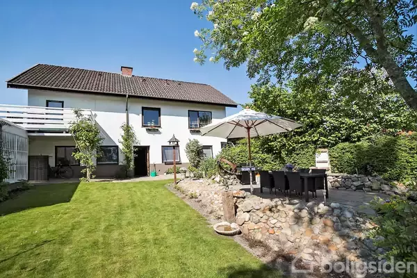 Hus med hvid facade og mørkt tag står i sollys, omkranset af grønne træer og buske. Terrassen har en parasol og udendørs siddepladser, mens haven har en velplejet græsplæne.