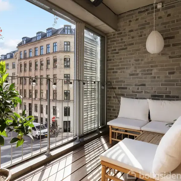 Balkon med hvid sofa og hængelampe. Placeret mod en murstensvæg, med udsigt over en gade med klassiske bygninger. Grøn plante til venstre.