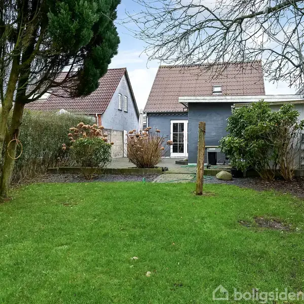 Huset med gråt eksteriør og rødt tegltag er omgivet af en have med grøn græsplæne, buske og træer. Der er en terrasse foran huset og et fuglehus i træet.